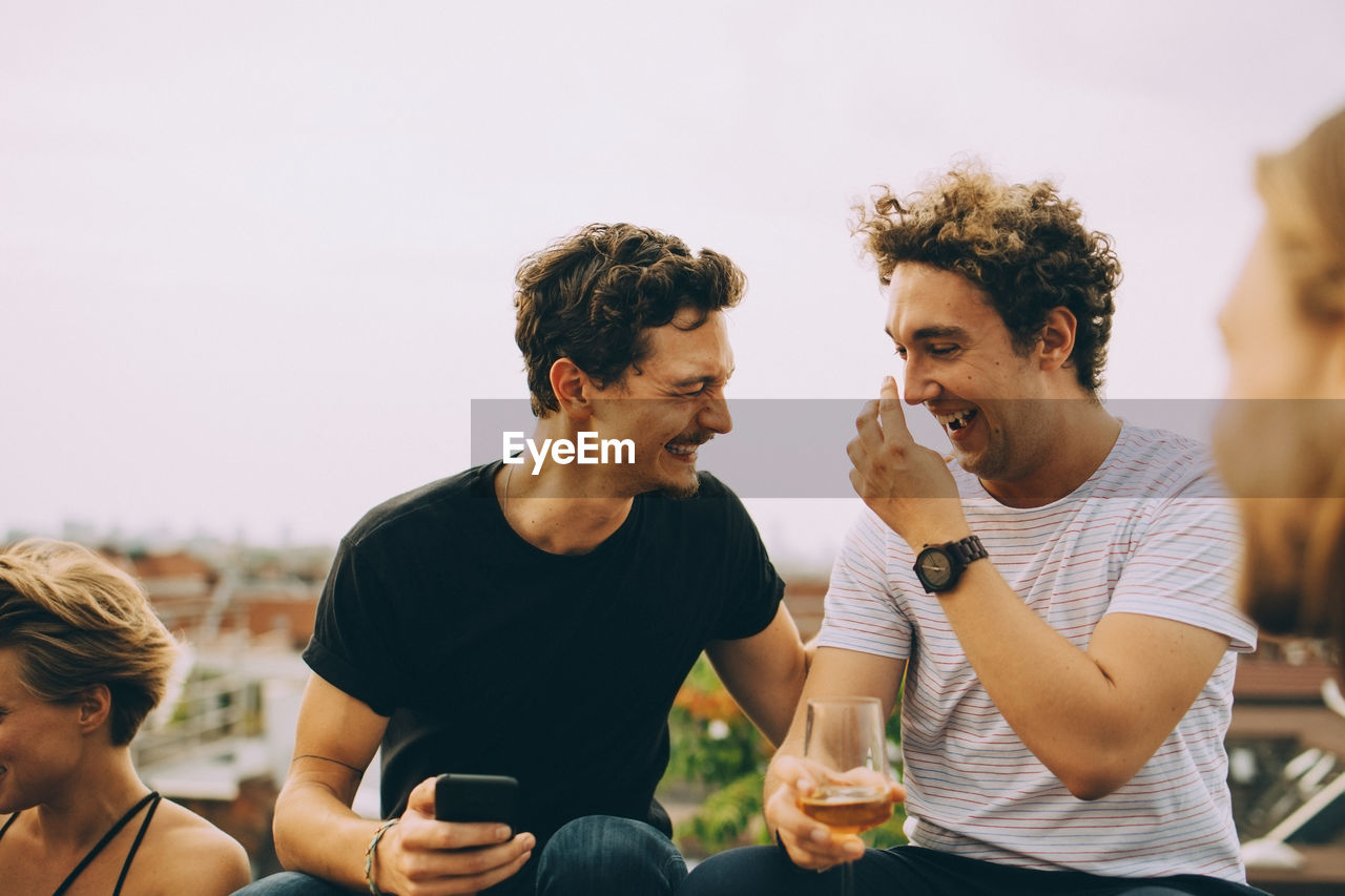 Happy man showing mobile phone to friend having drink at terrace during party