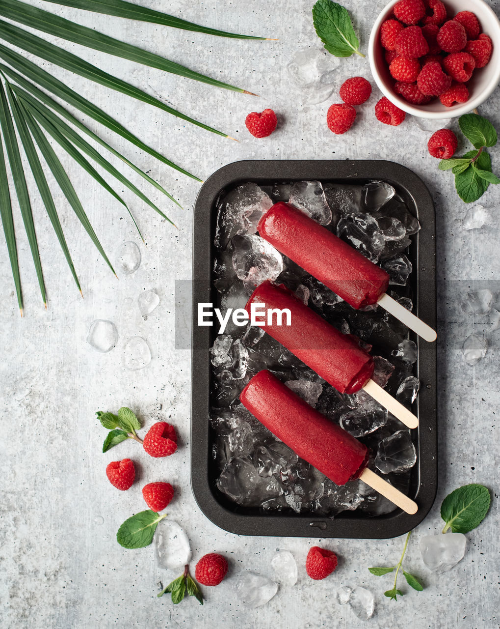 Raspberry popsicles on a tray with ice and fruit on grey background.