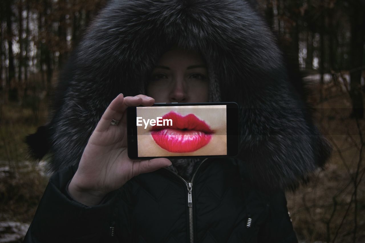 Portrait of young woman holding smart phone showing pink lips in display during winter