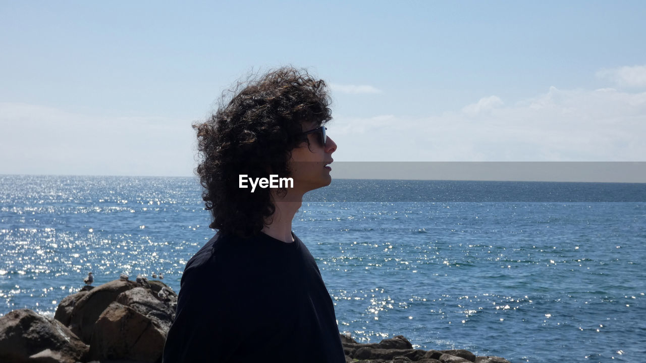 Side view of young man wearing sunglasses looking at sea while standing against sky during sunny day