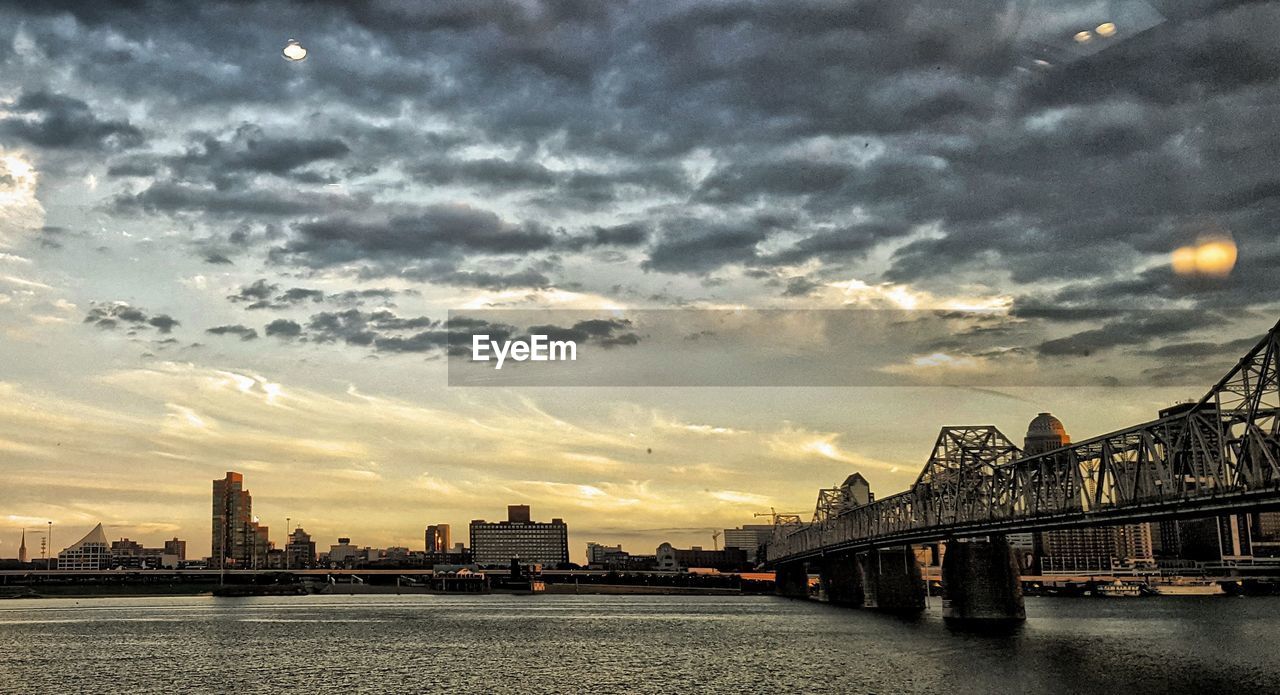 BRIDGE OVER RIVER AGAINST CLOUDY SKY IN CITY