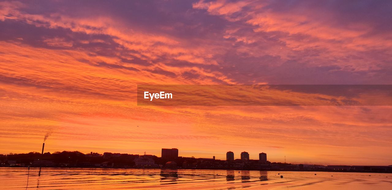 SILHOUETTE BUILDINGS BY SEA AGAINST ROMANTIC SKY AT SUNSET