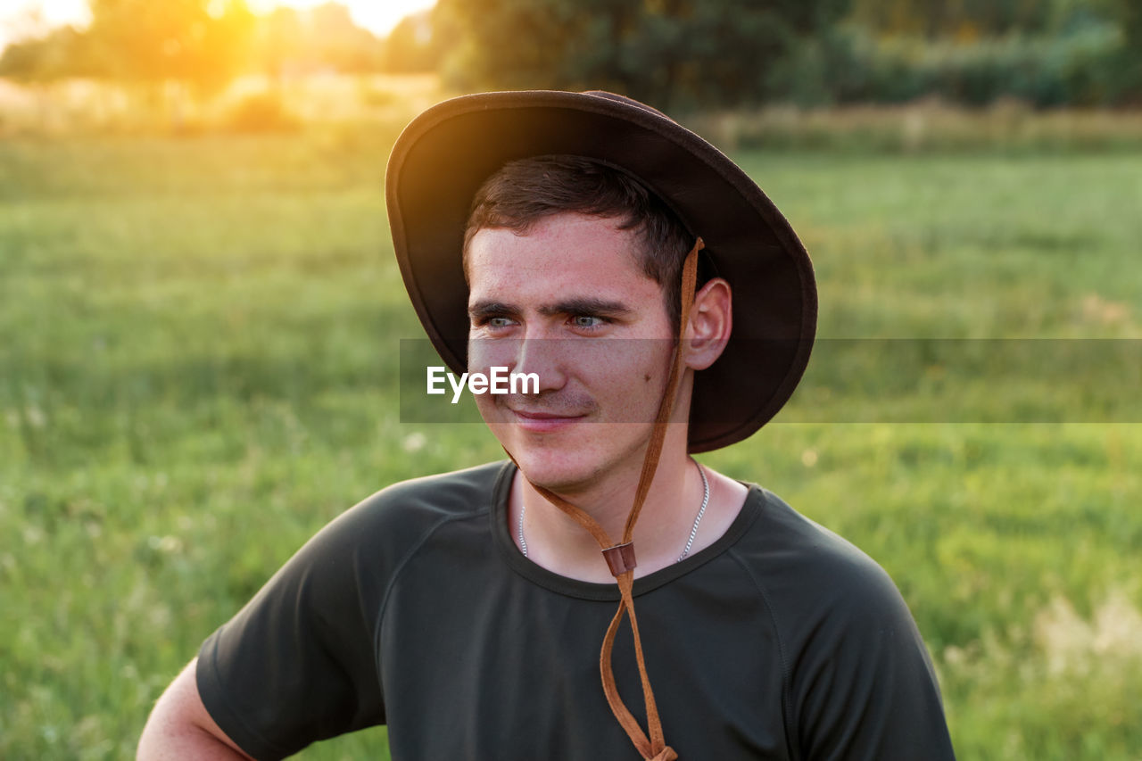 Young man smiling farmer in cowboy hat at agricultural field on sunset with sun flare. closeup 