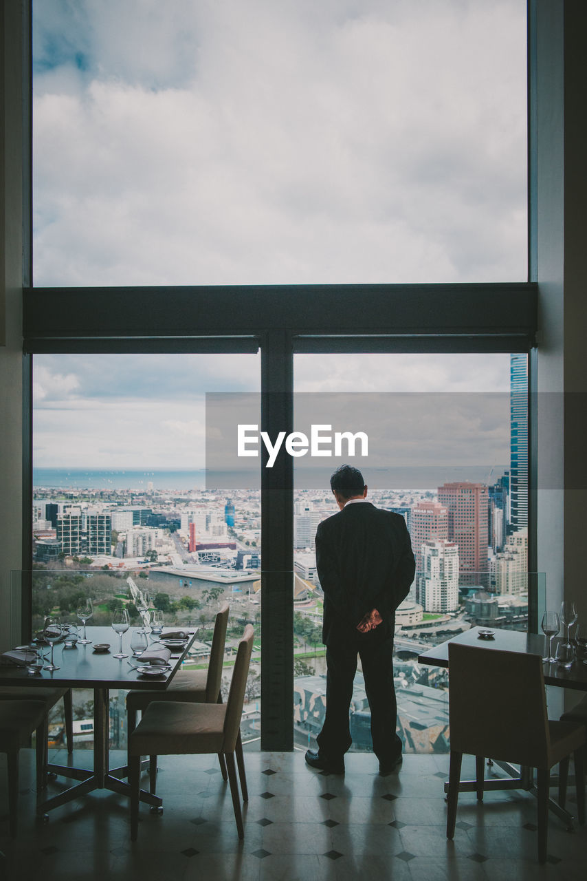 REAR VIEW OF MAN STANDING BY WINDOW WITH CITYSCAPE