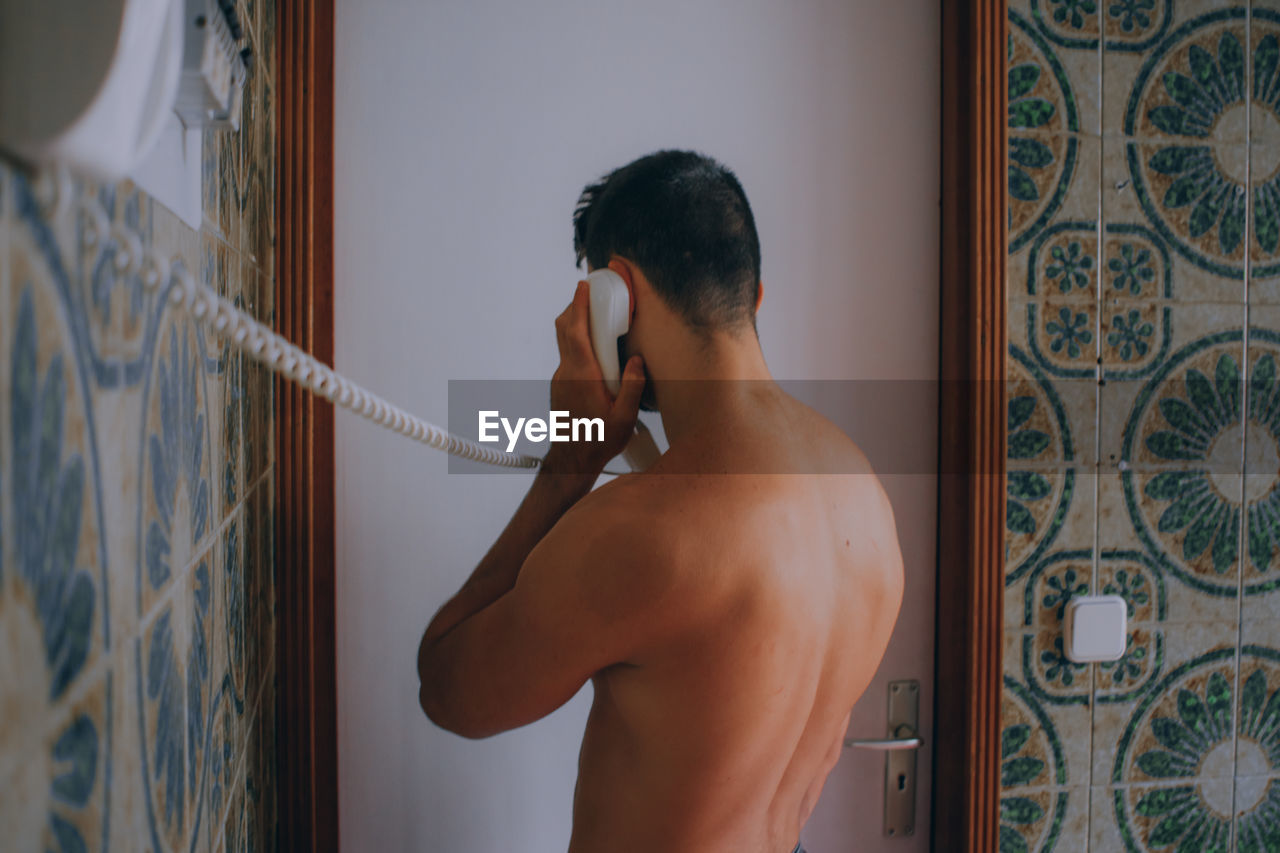 Rear view of shirtless man talking on phone while standing against wall at home