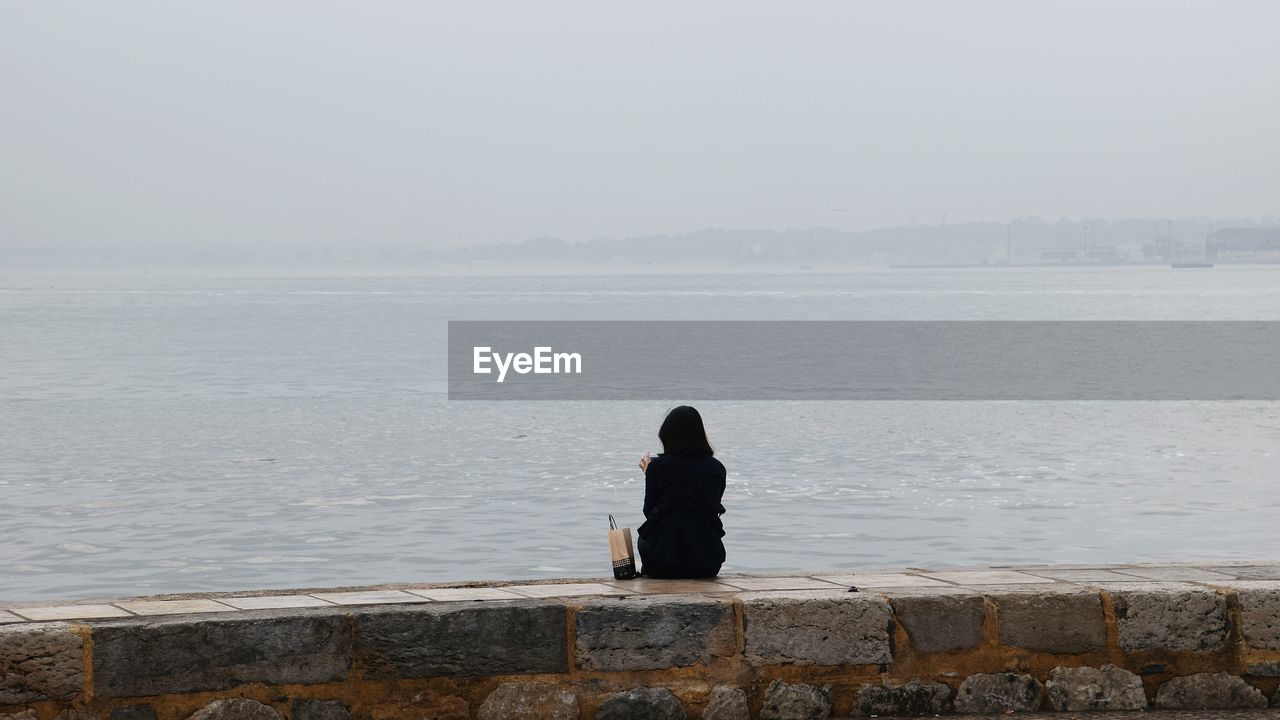 Rear view of woman sitting on retaining wall by sea against clear sky