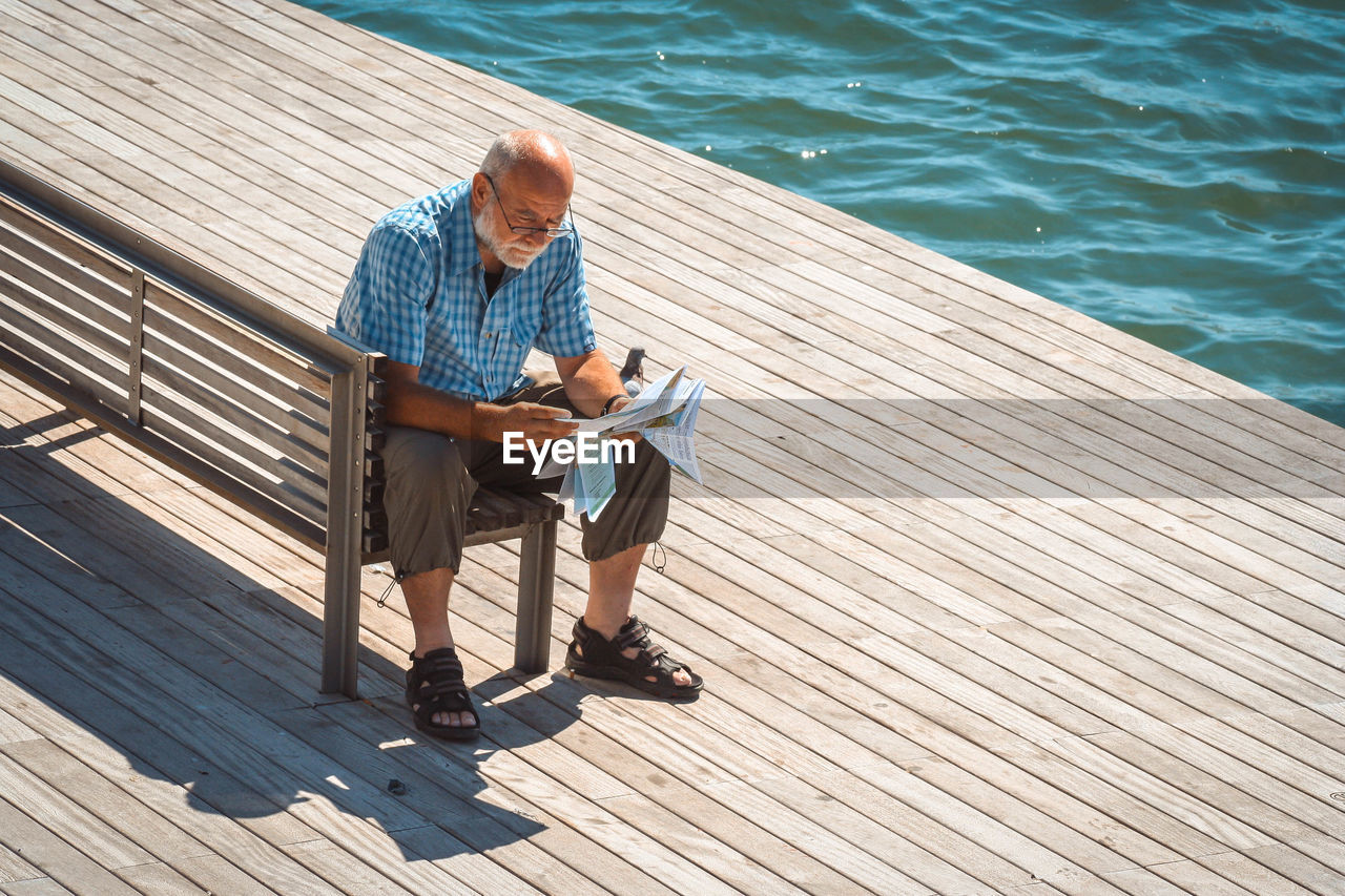 Full length of man reading paper while sitting by lake on bench during sunny day