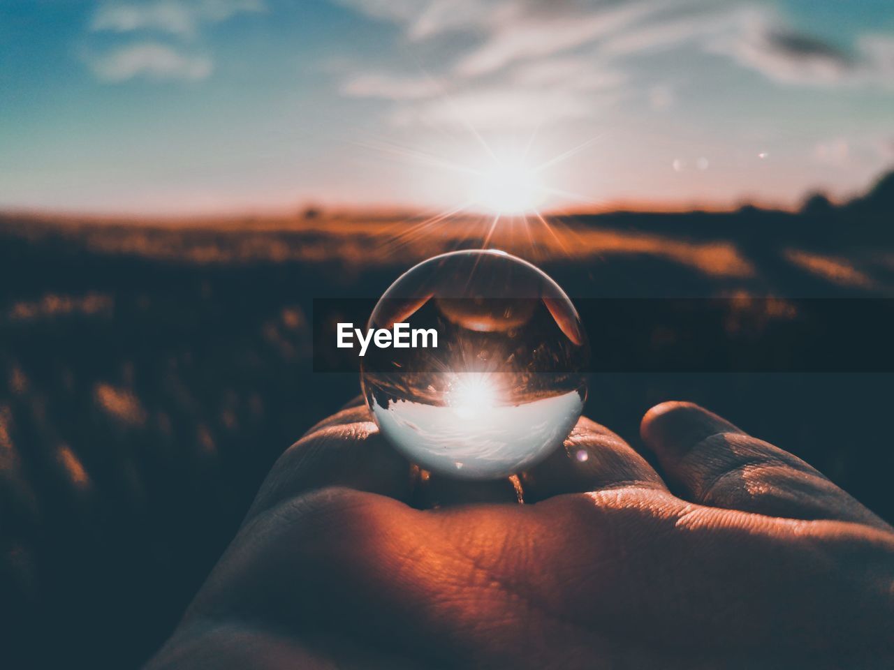 Cropped hand of person holding crystal ball against sky during sunset