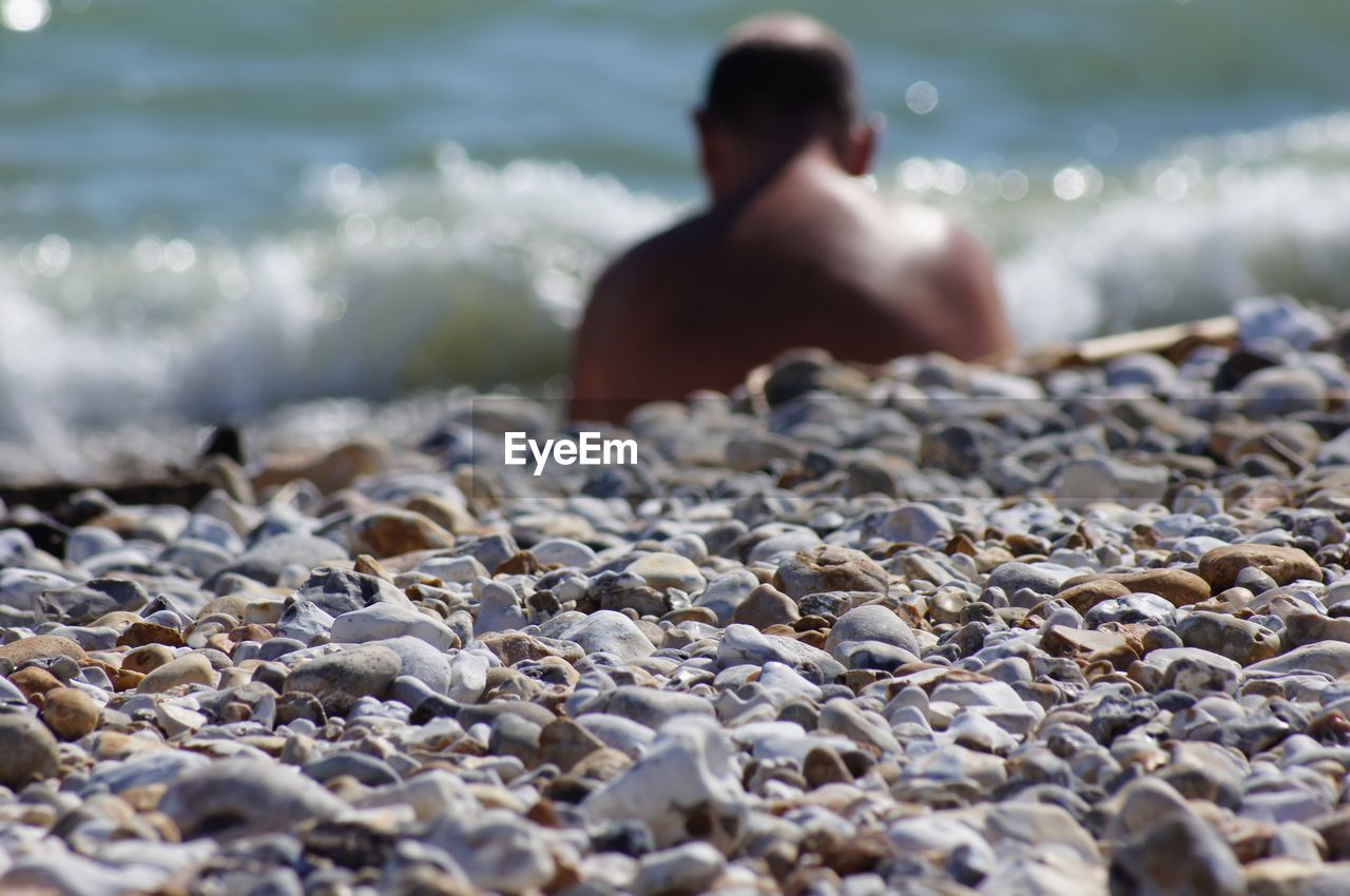 REAR VIEW OF SHIRTLESS MAN LYING ON PEBBLES AT BEACH