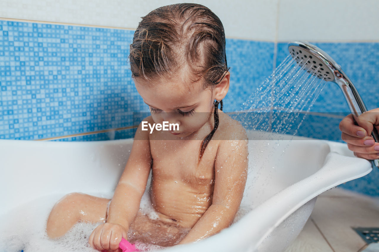 Cropped hand of mother showering shirtless daughter sitting in bathtub
