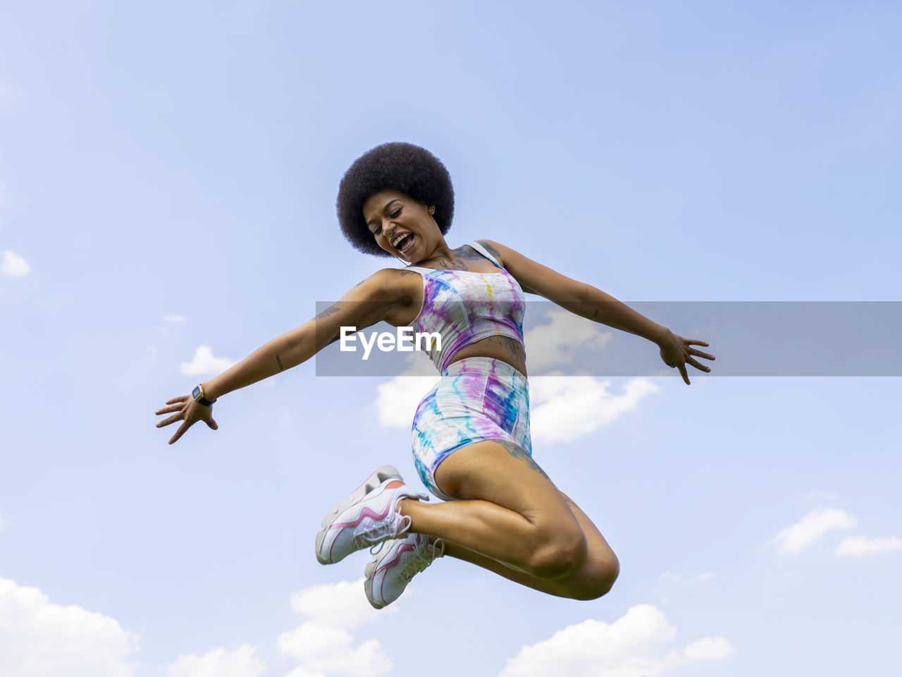 Low angle of delighted african american female in summer outfit in moment of jumping above ground on background of blue sky