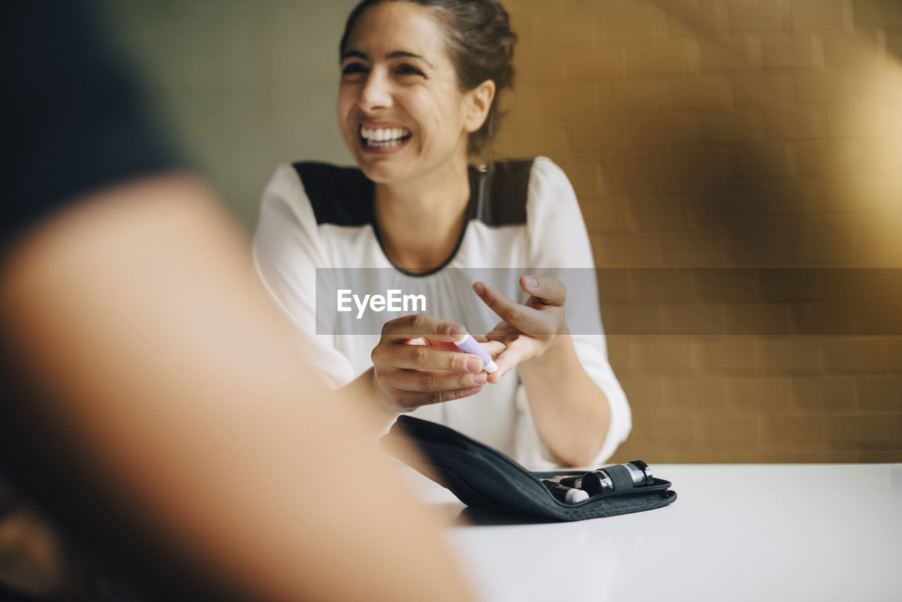 Smiling businesswoman doing blood sugar test while sitting with colleague at table in office