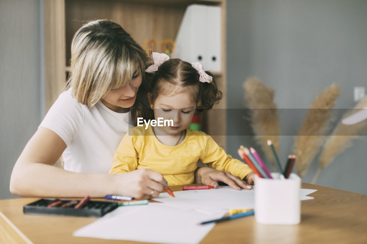 Happy mother and daughter draw together with colored pencils at the table in the room. time together