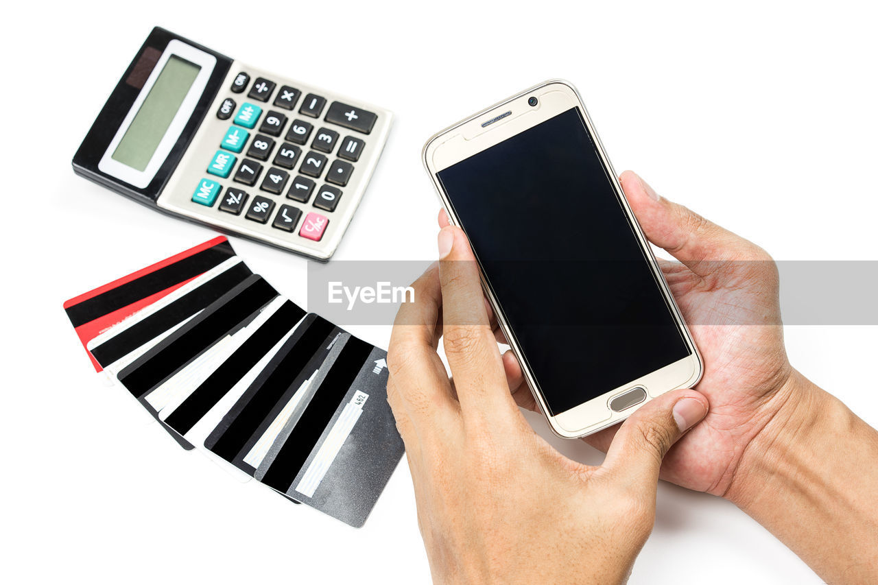 Cropped hands of person holding smart phone by credit cards and calculator against white background