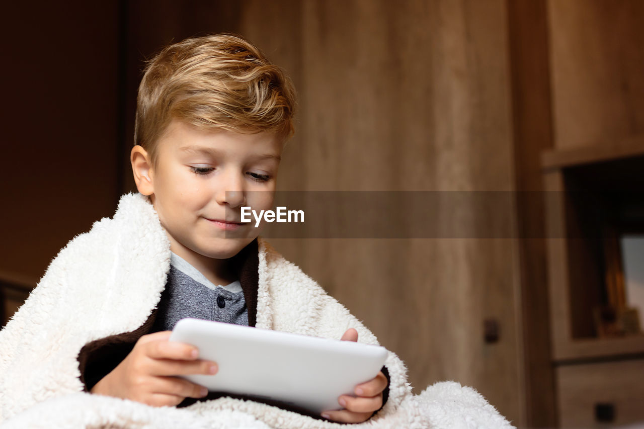 Small boy wrapped in a blanket using digital tablet at home.