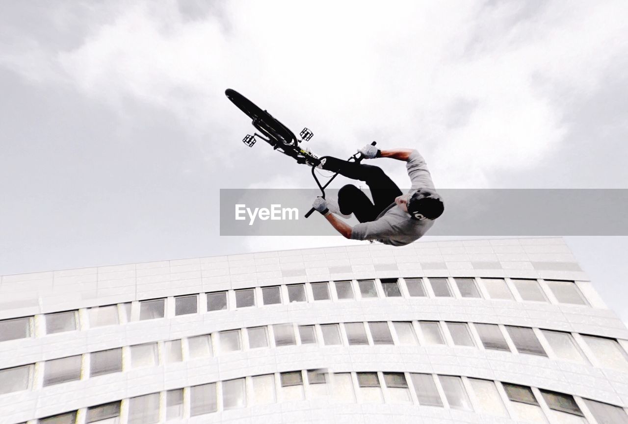 Low angle view of man doing stunt with bicycle by building against sky