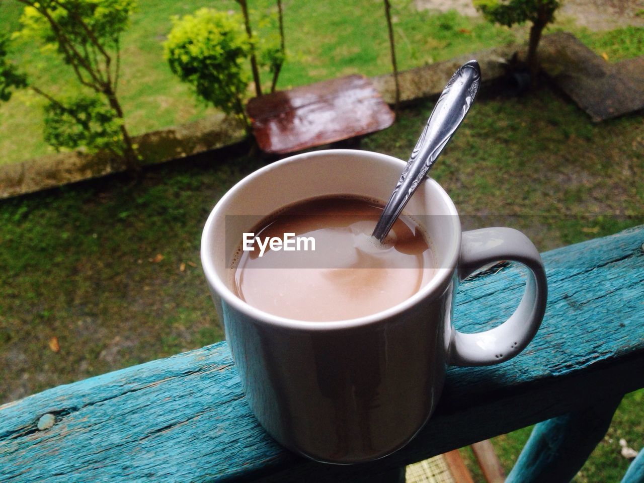 Close-up of coffee cup with spoon on wooden railing by lawn