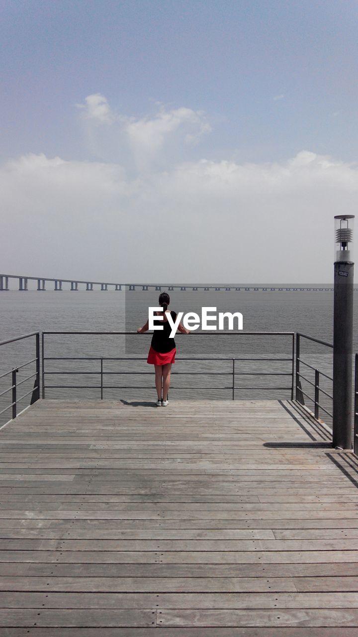 REAR VIEW OF WOMAN STANDING ON PIER AGAINST SEA