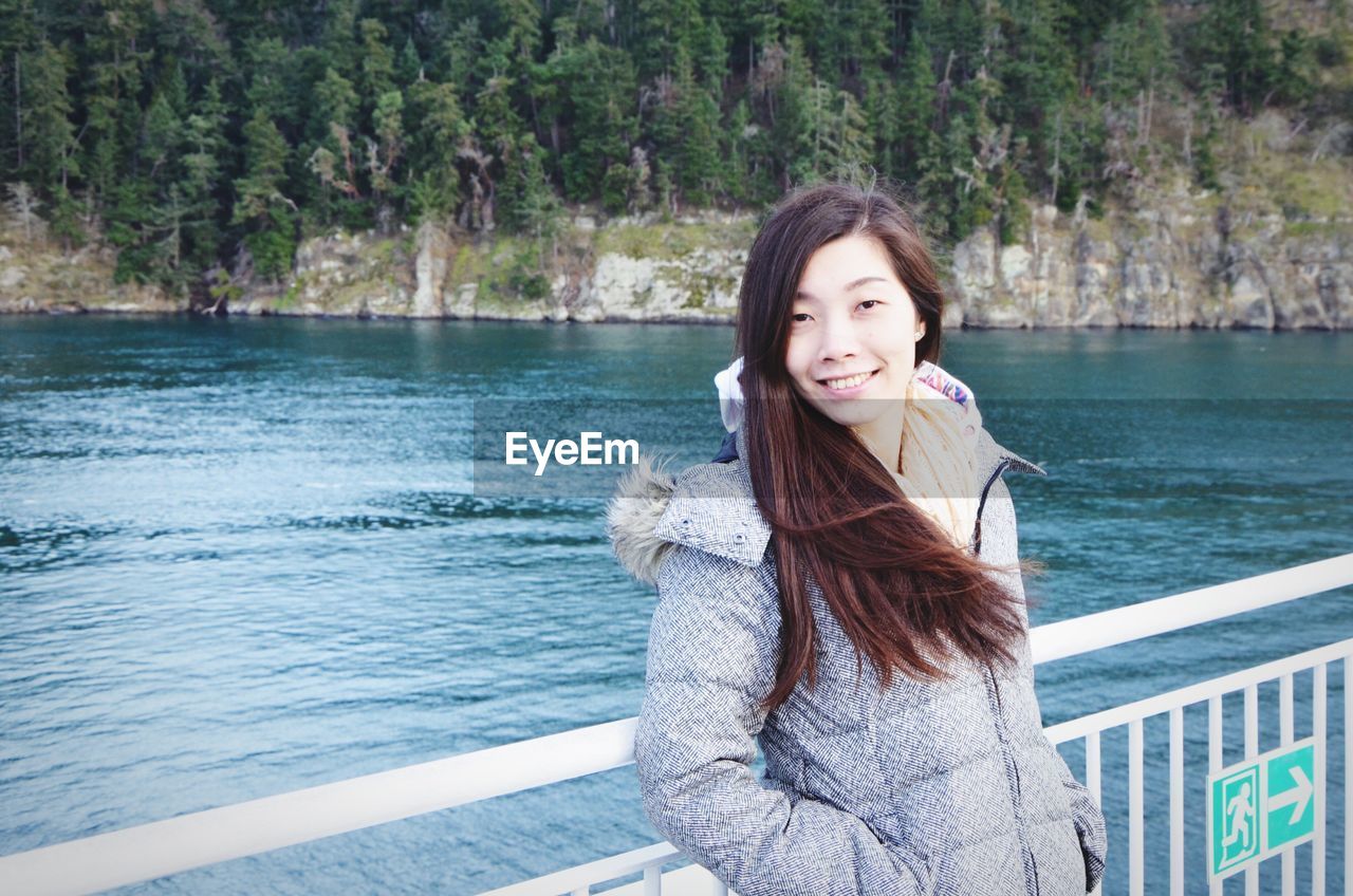 Portrait of smiling woman standing at railing by lake