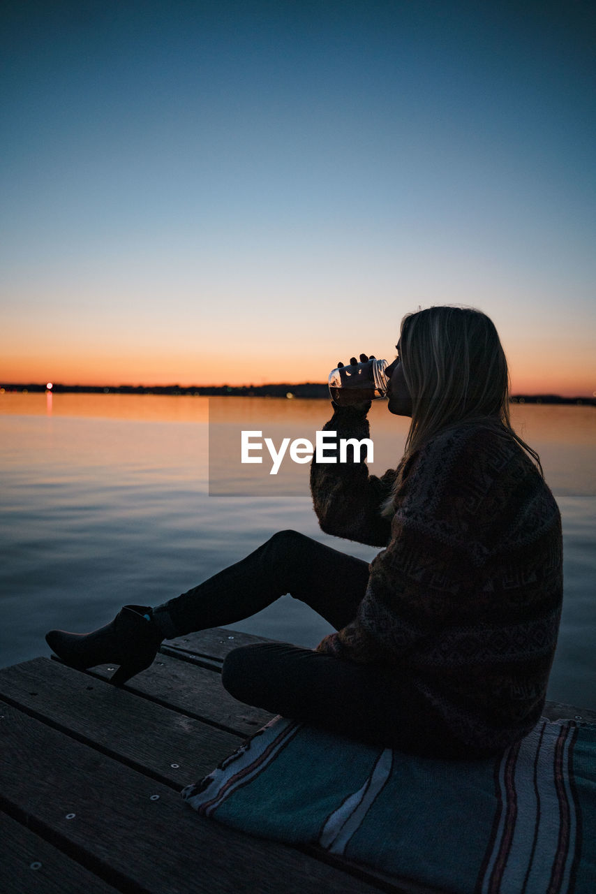 Side view of woman having drink while sitting on pier over lake against clear sky during sunset