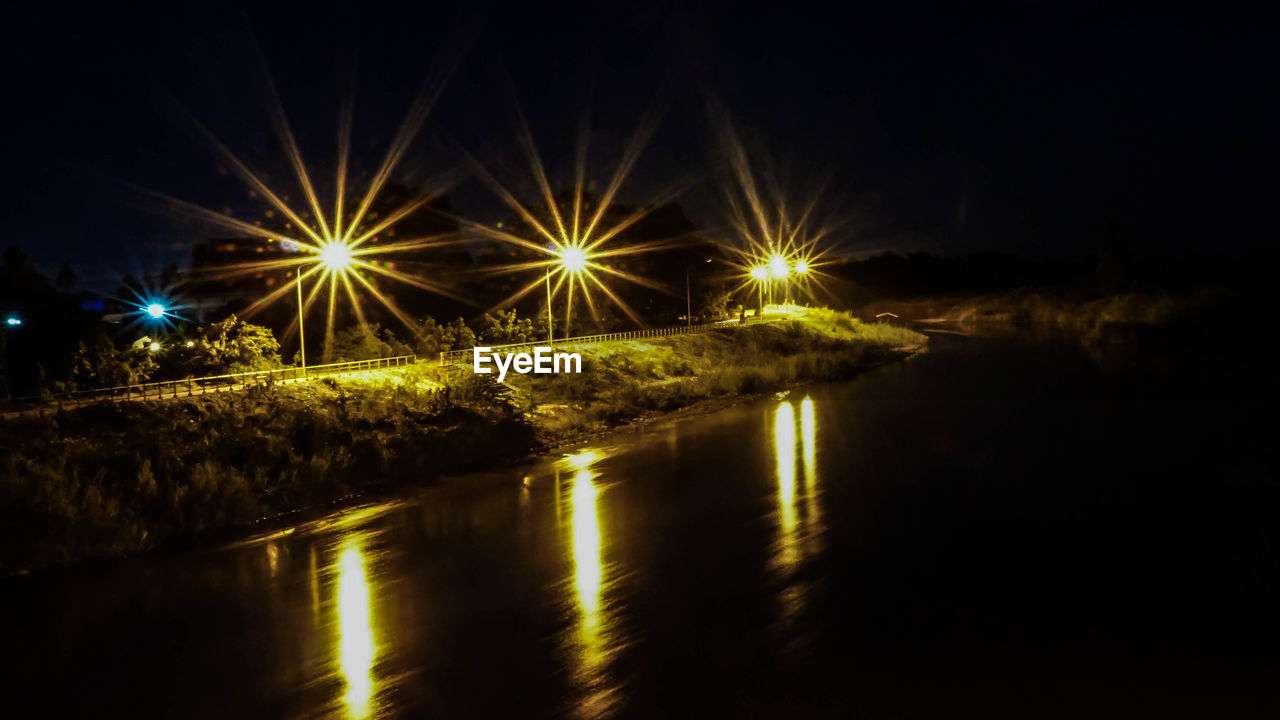 Illuminated street lights by river against sky at night