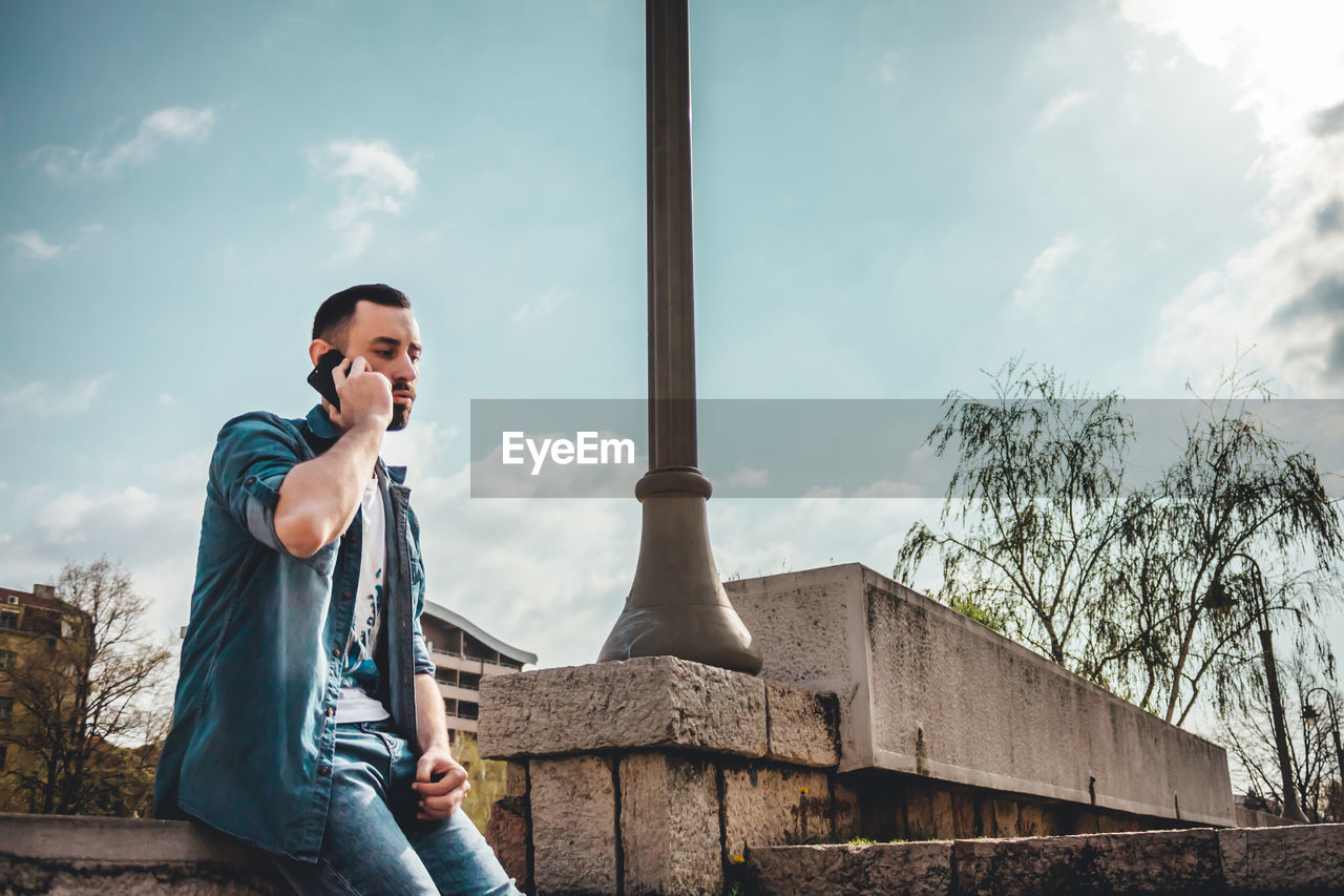 YOUNG MAN USING MOBILE PHONE WHILE SITTING ON SKY