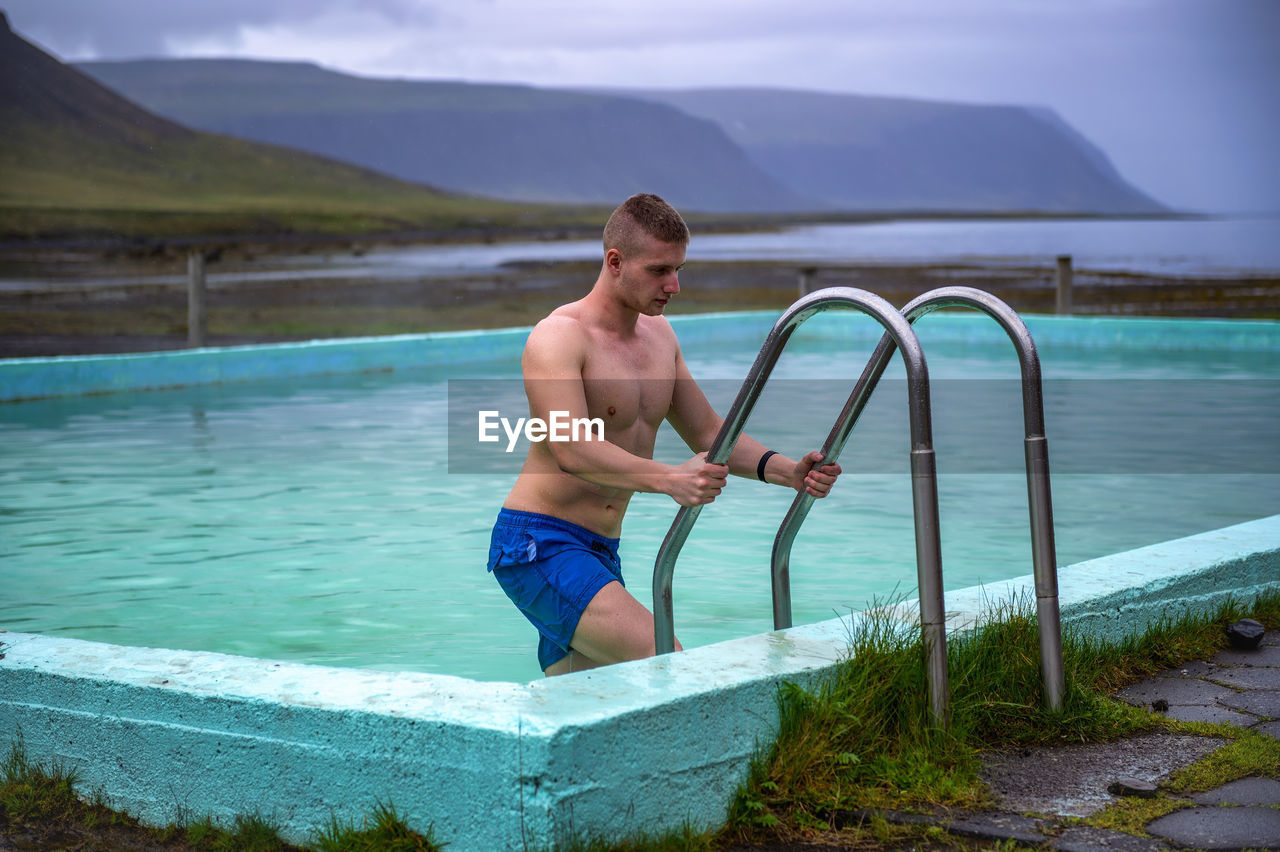 FULL LENGTH OF SHIRTLESS MAN STANDING BY SWIMMING POOL