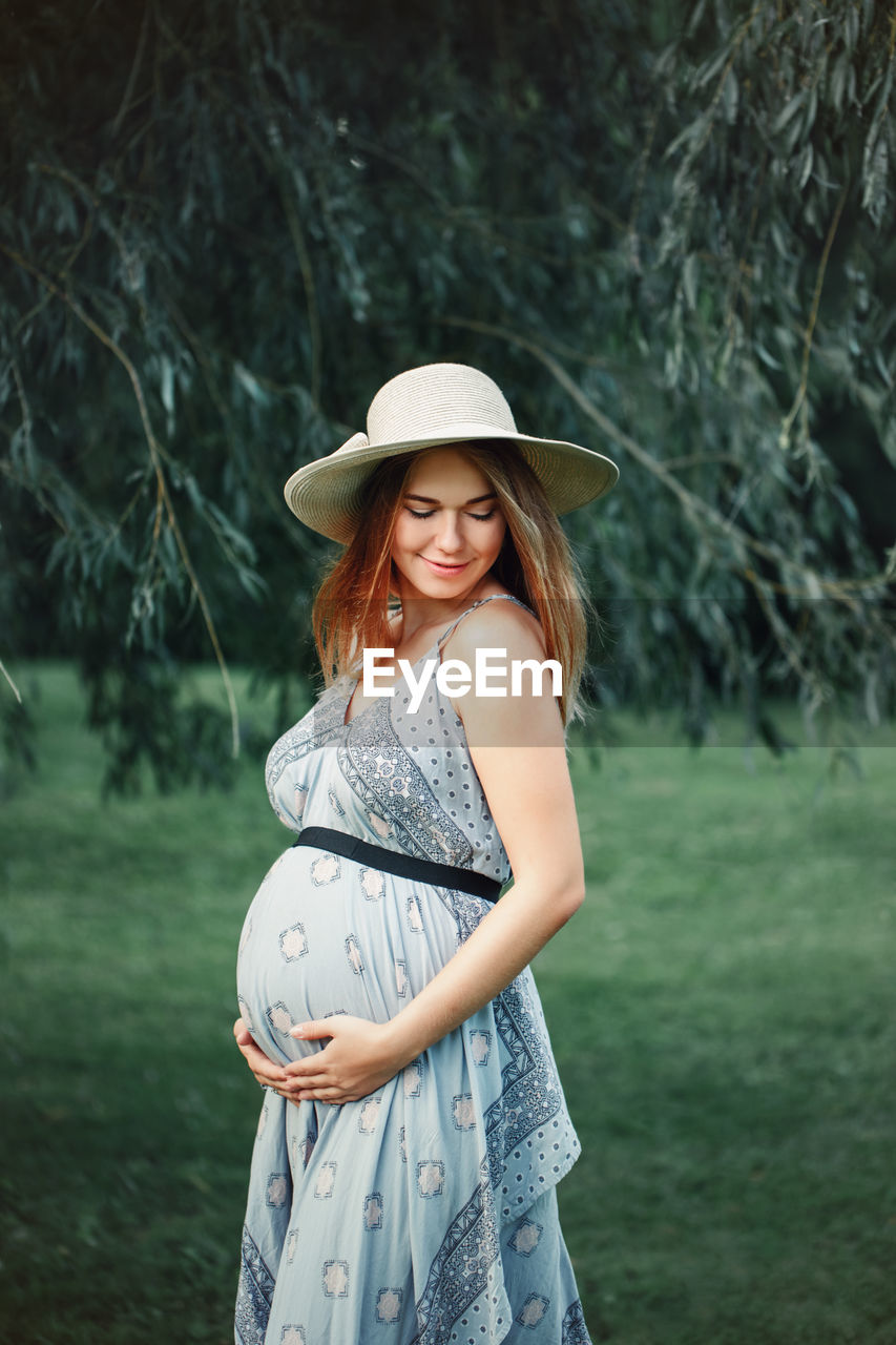 Smiling pregnant woman touching abdomen while standing on grass