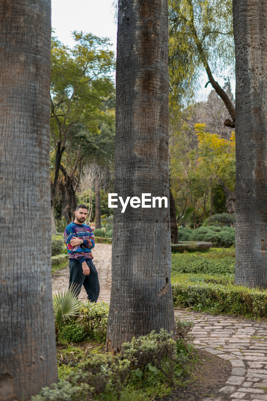 Caucasian model standing in between palm trees in a public park