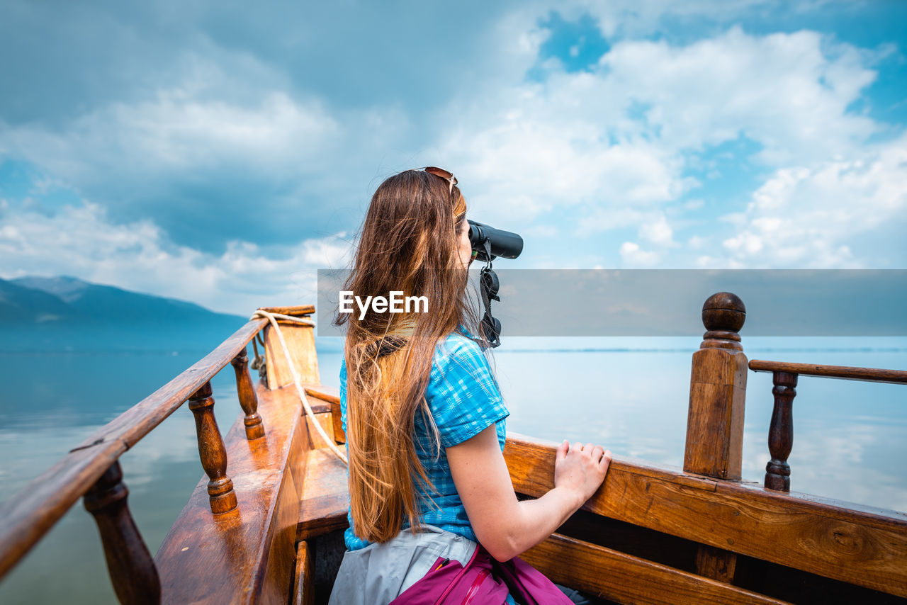 Side view of woman looking through binoculars while sitting in boat against sky