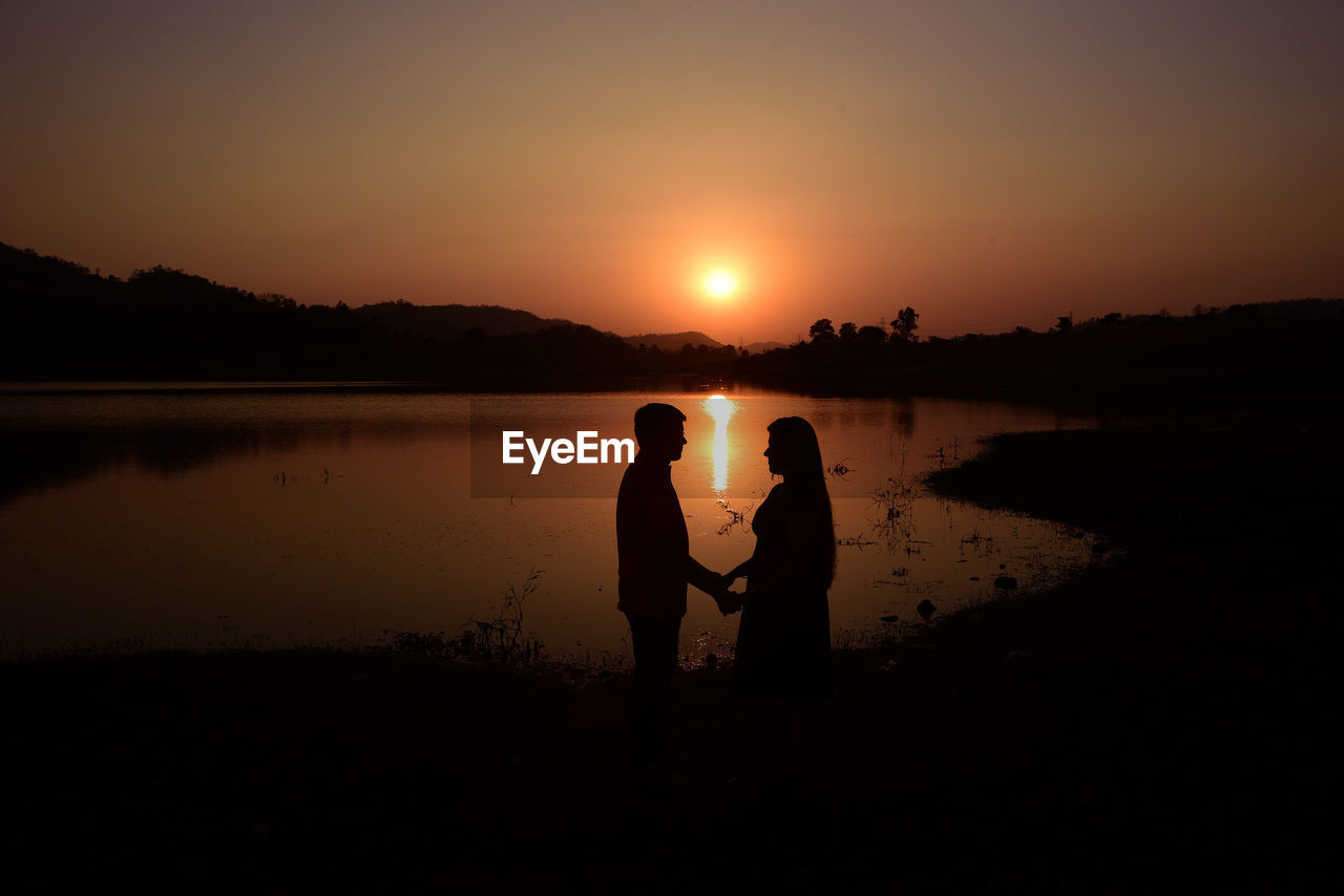 Silhouette boy and girl' standing on shore against sky during sunset