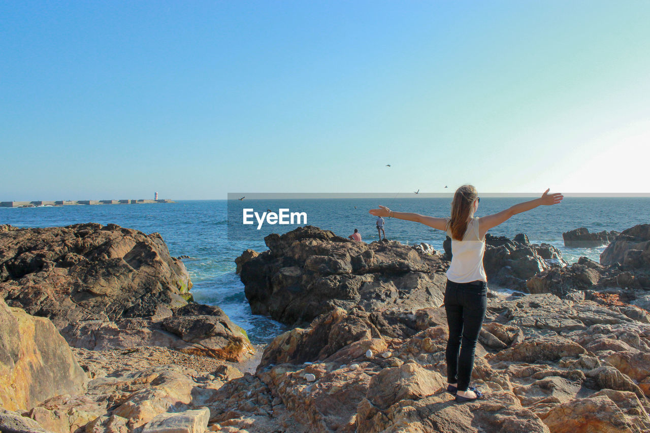 Rear view of woman standing on rocks against sea and clear sky