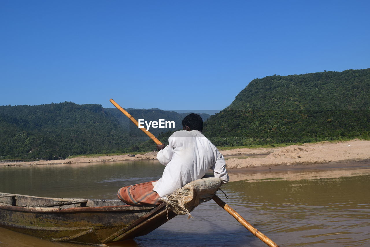 Rear view of man rowing boat in lake against clear blue sky