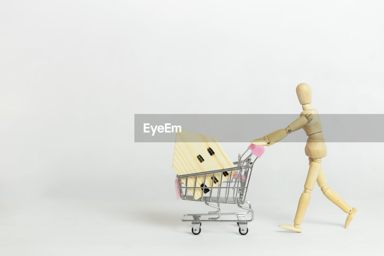 Wooden mannequin or puppet pushing shopping cart with wooden house on white background.