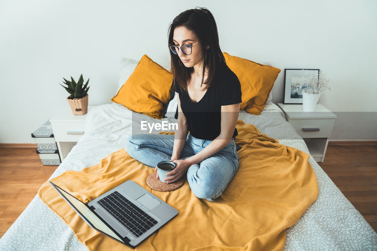 Young woman relaxing and holding cup of hot coffee. female sitting on bed and surfing the net