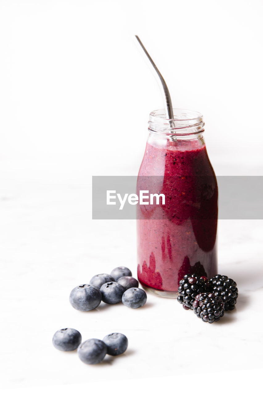 Smoothie in a glass bottle, metal straw, blueberries and blackberries on a marble surface. front shot.