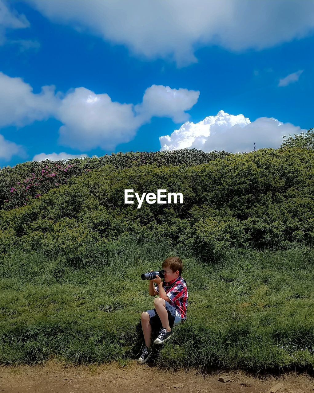 Side view of boy photographing with camera while sitting on grassy field