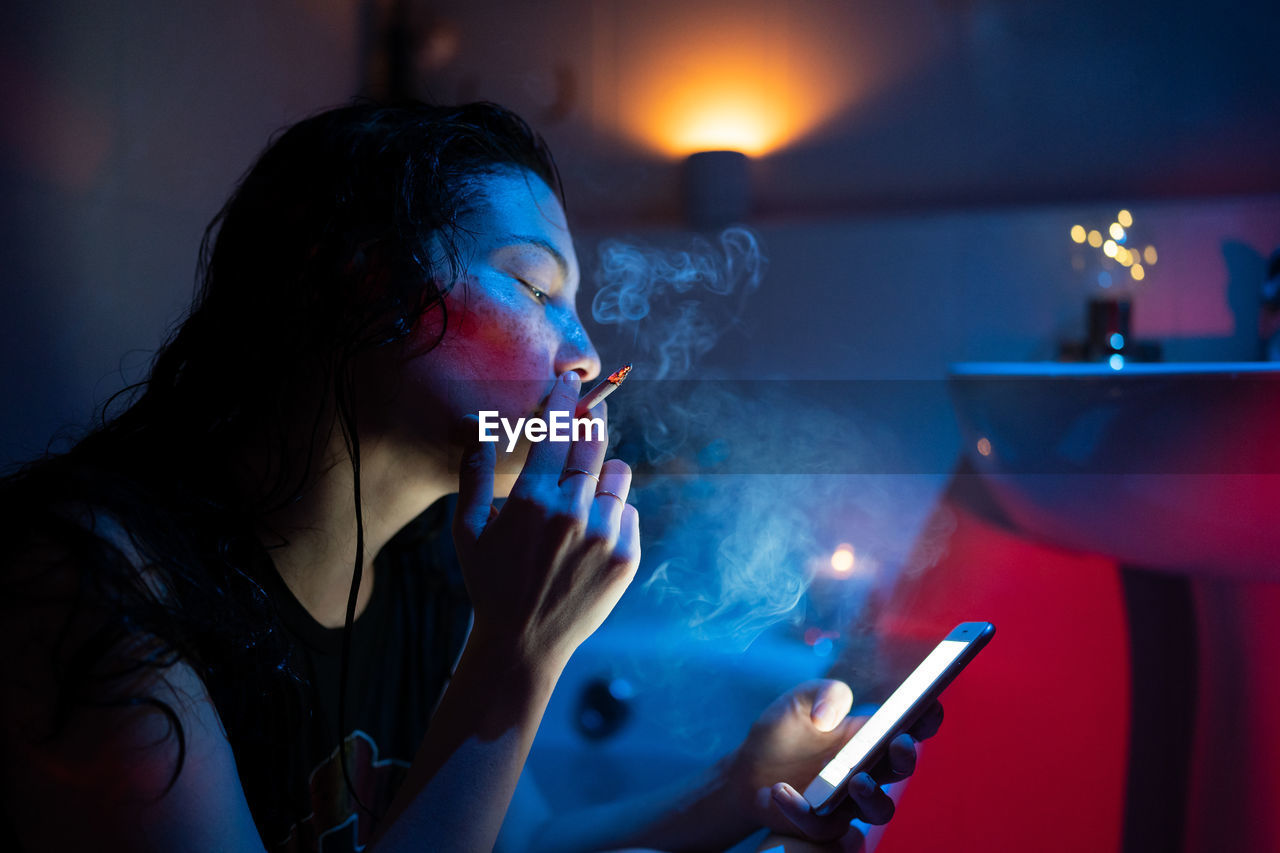 Young woman addicted to nicotine smoking cigarette and using smartphone under neon light in bathroom