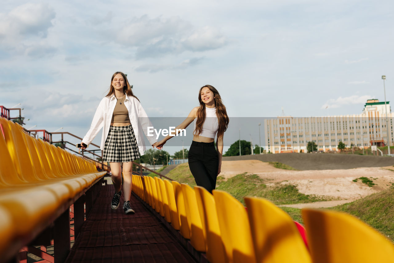 Two teenage girls walk together through the stands of the school stadium, talking, holding hands, 