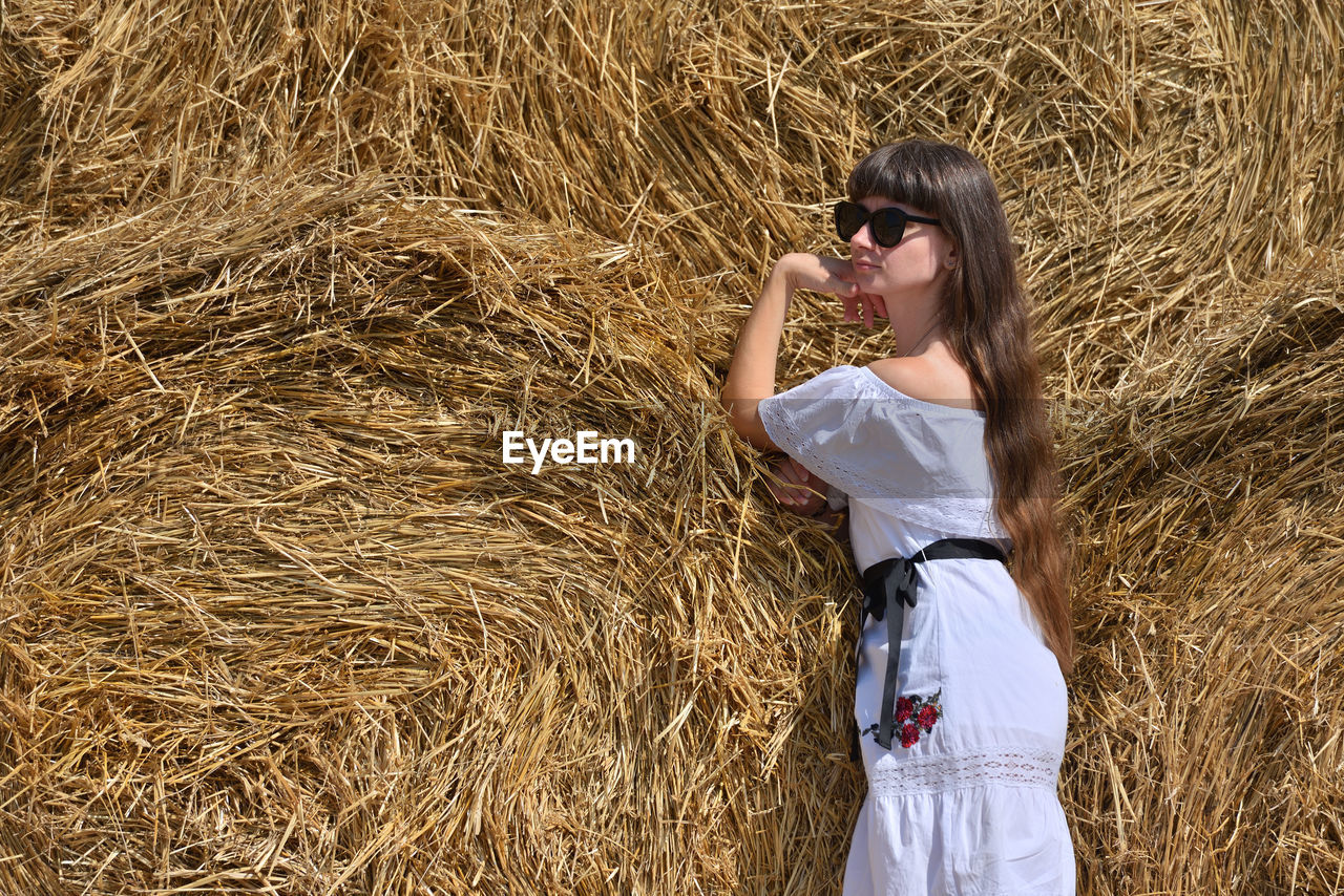 Mid adult woman standing against stacked hay bales on agricultural field