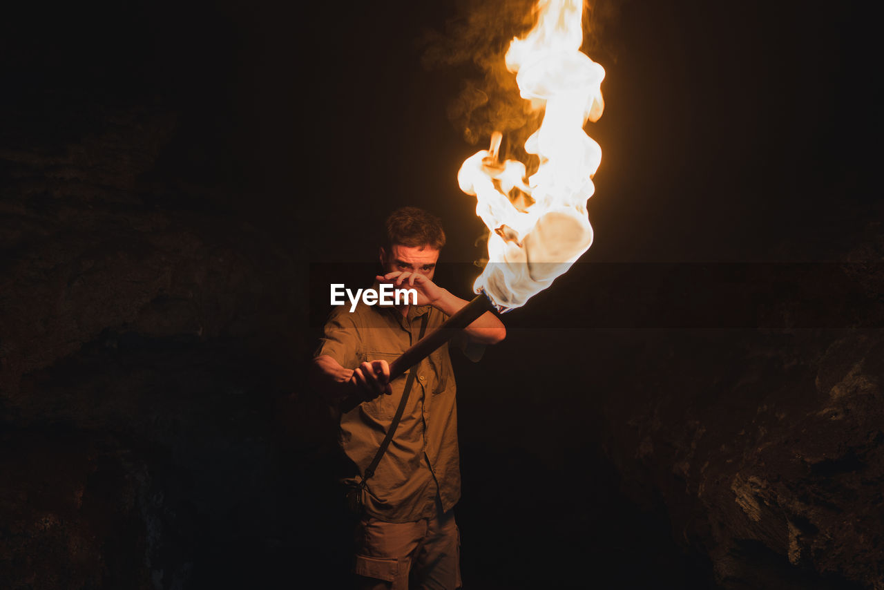 Young male speleologist with flaming torch standing in dark narrow rocky cave while exploring subterranean environment