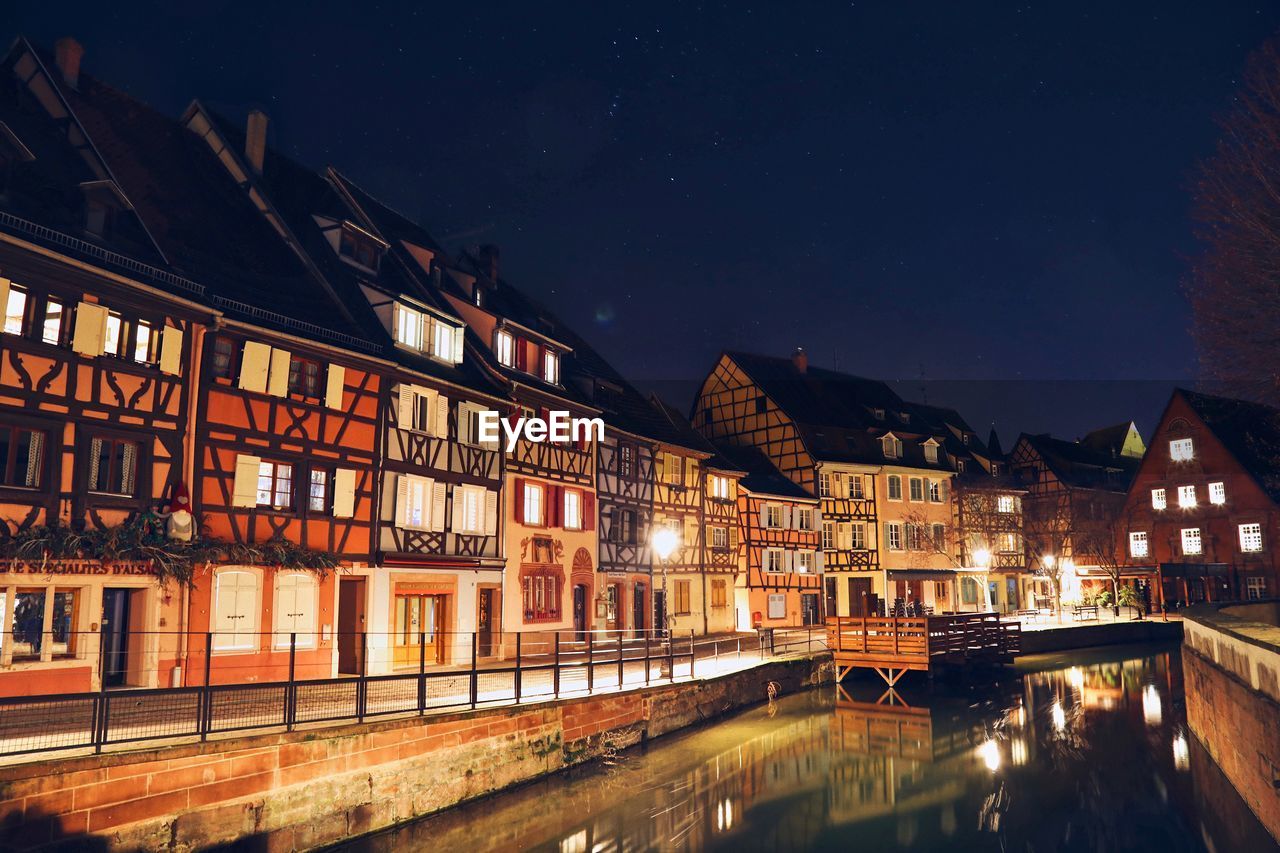 ILLUMINATED BUILDINGS BY RIVER AGAINST SKY AT NIGHT