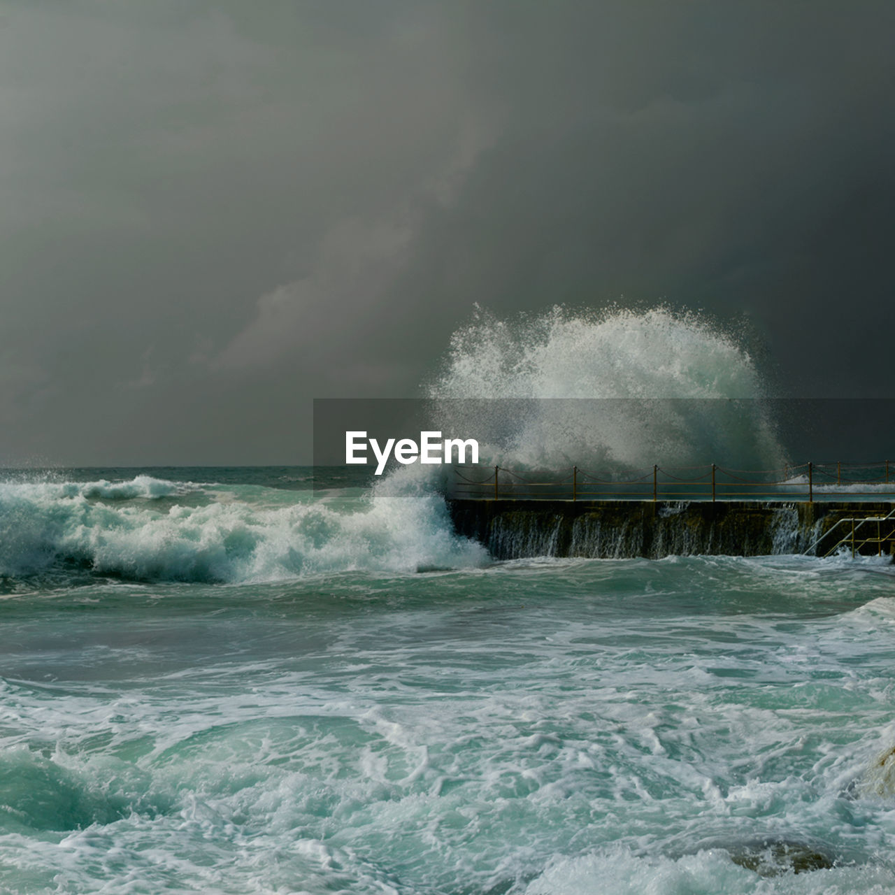 Sea waves splashing on pier against cloudy sky