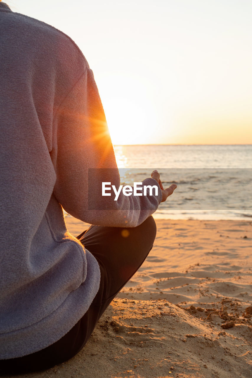 Midsection of man sitting at beach doing yoga on sunrise 