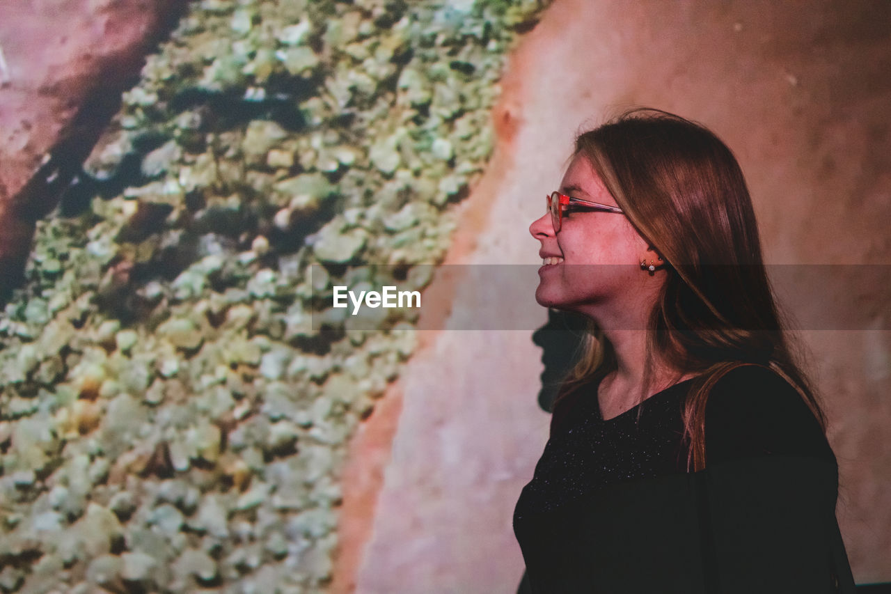 Smiling woman looking away while standing against wall at night