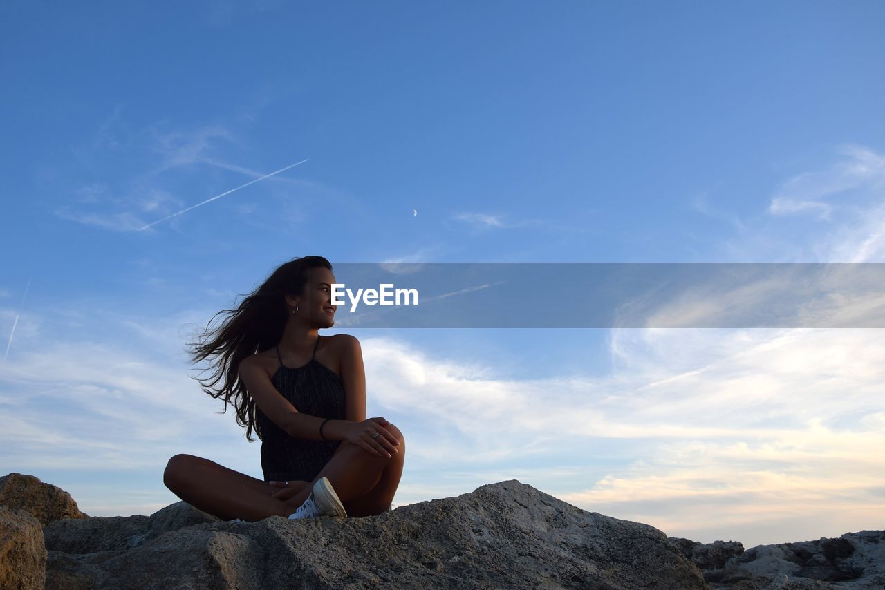 Young woman sitting on rock against sky