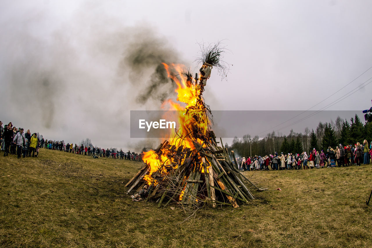 Crowd gathered by scarecrow burning on field against sky