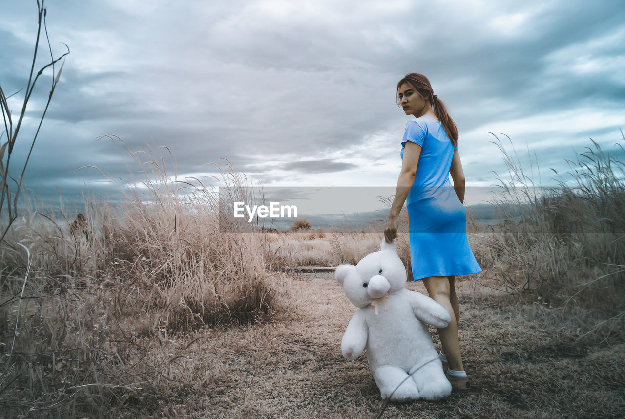 Full length of woman standing with teddy bear on field against cloudy sky