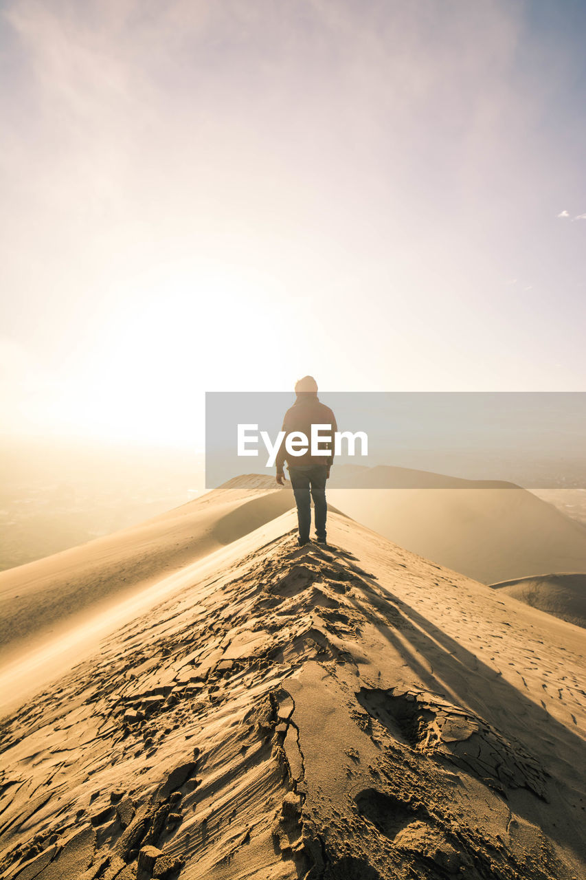 Rear view of man standing on sand dune against sky during sunset