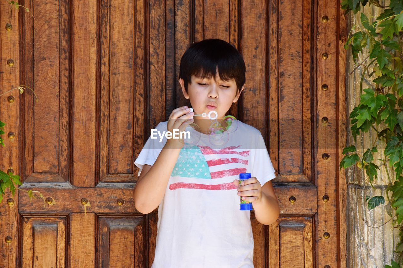 Cute boy blowing bubbles while standing against wood